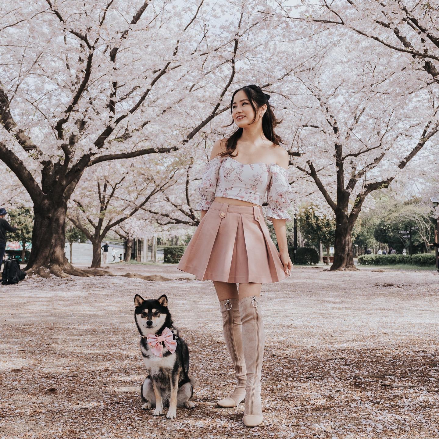 A woman poses with a dog in a park with blossoming trees. She wears a pink skirt, a floral top, and high boots. The dog has a pink bow.