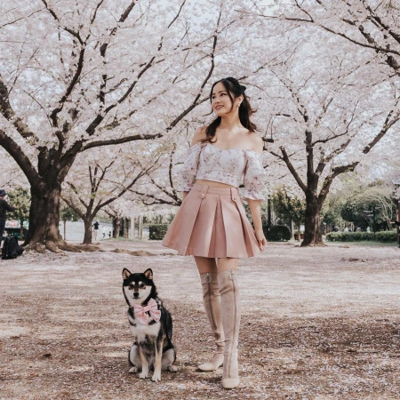 A woman poses with a dog in a park with blossoming trees. She wears a pink skirt, a floral top, and high boots. The dog has a pink bow.