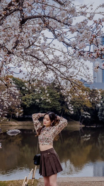 Young woman in a floral top and brown skirt poses near a pond surrounded by cherry blossom trees in a park setting on a cloudy day.