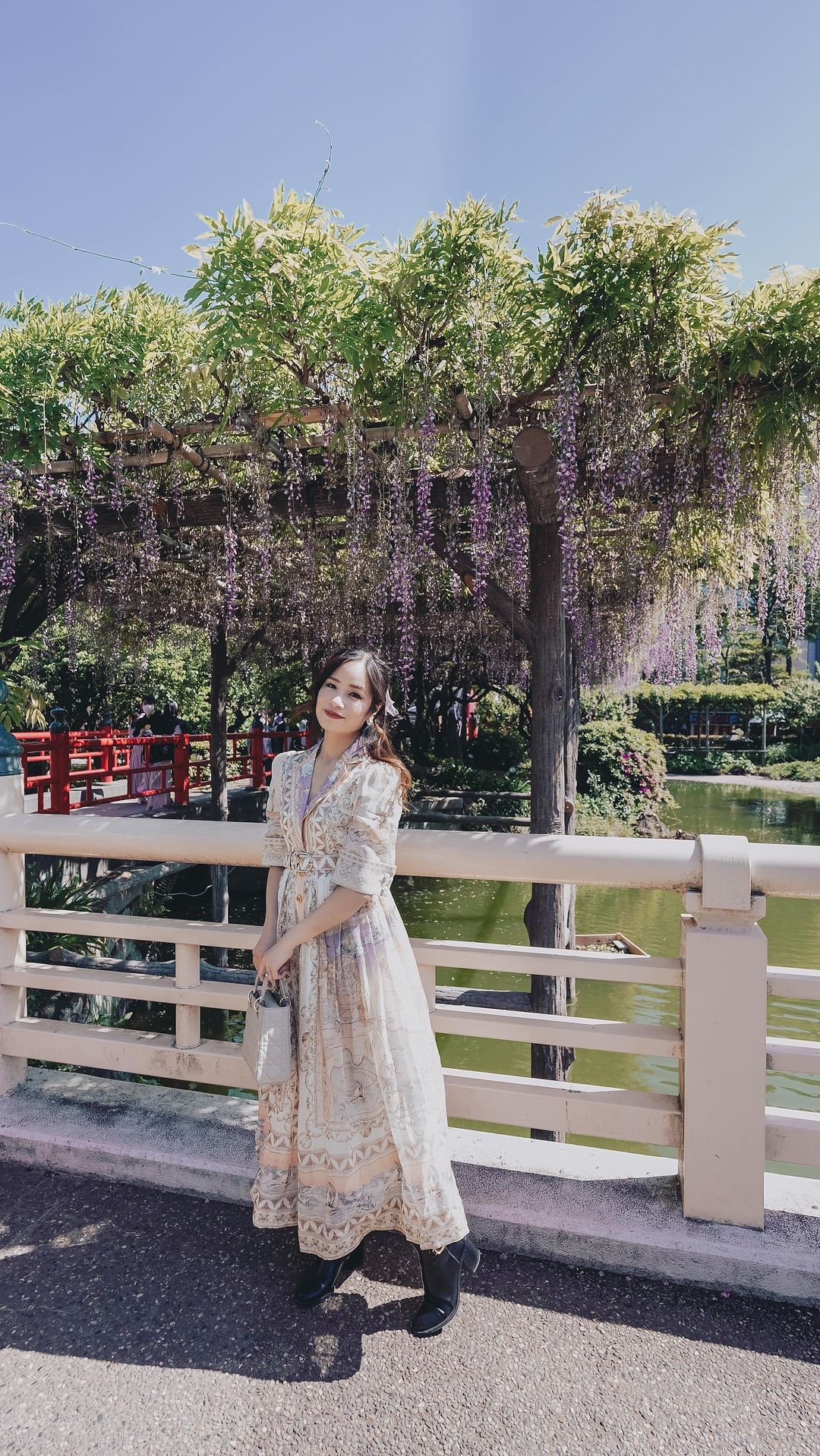A woman in a long floral dress poses on a bridge with a wisteria-covered arbor in the background on a sunny day.