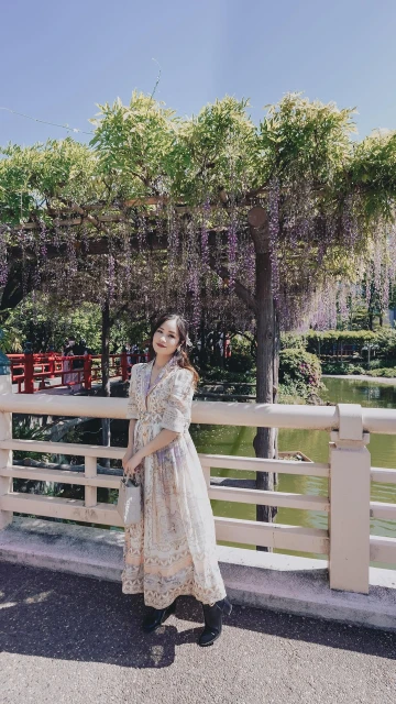 A woman in a long floral dress poses on a bridge with a wisteria-covered arbor in the background on a sunny day.
