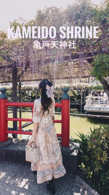 Woman in a patterned dress gazes at the Kameido Shrine sign under a wisteria trellis with a red railing in an outdoor setting.