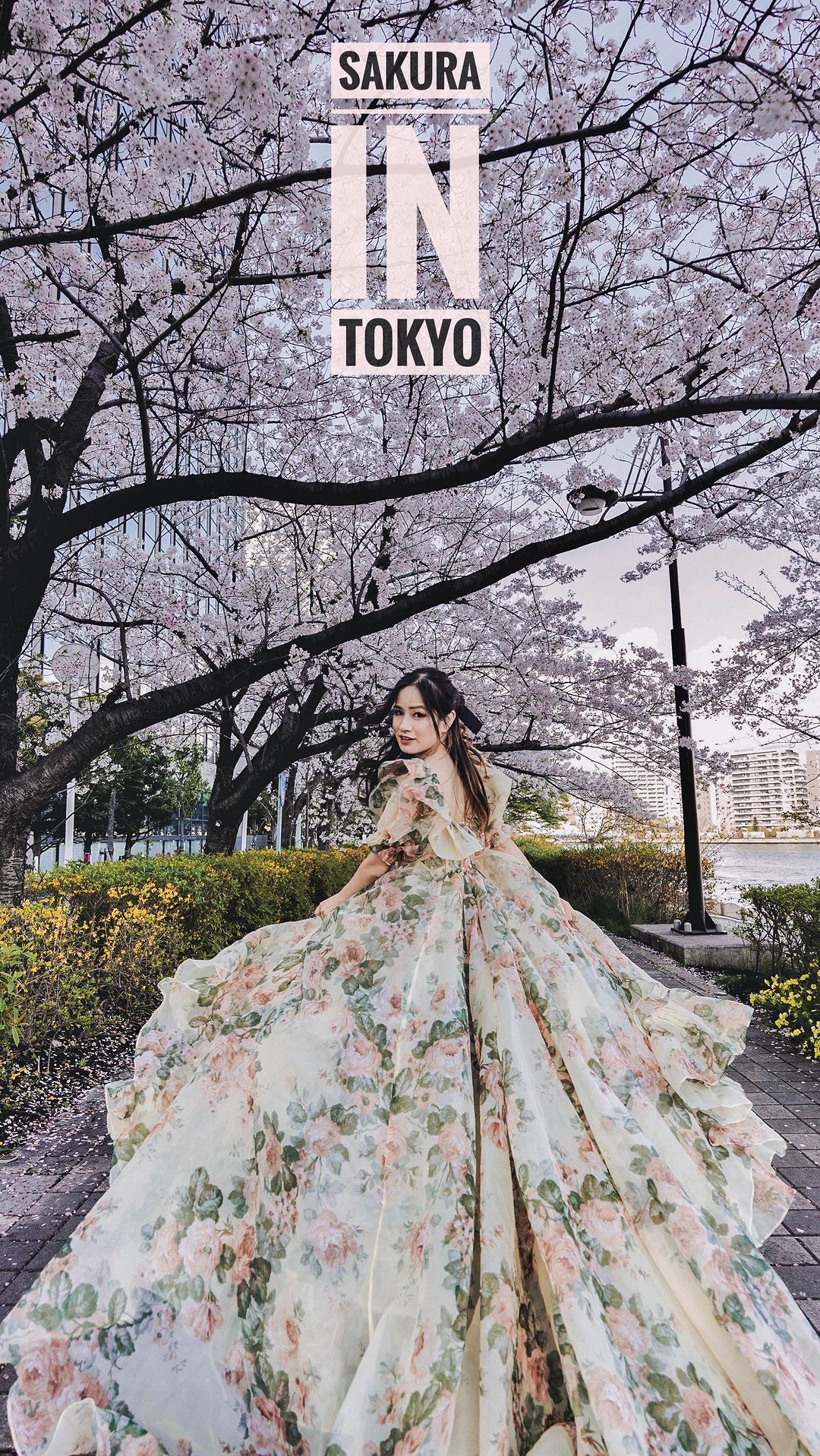 Woman in floral gown stands under cherry blossom trees in Tokyo, showcasing a blend of nature, fashion, and Japanese urban landscape.