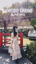 Woman in patterned midi dress holding a handbag standing before a red bridge under wisteria blossoms at Kameido Shrine.