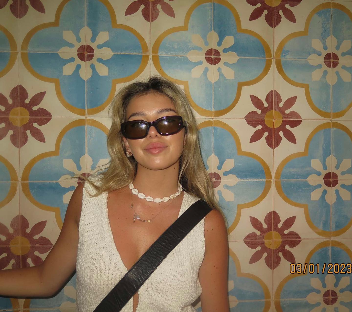 Indoor portrait of a young woman with long blonde hair and sunglasses standing in front of a colourful tiled wall.