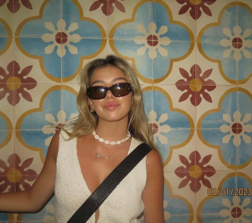 Indoor portrait of a young woman with long blonde hair and sunglasses standing in front of a colourful tiled wall.