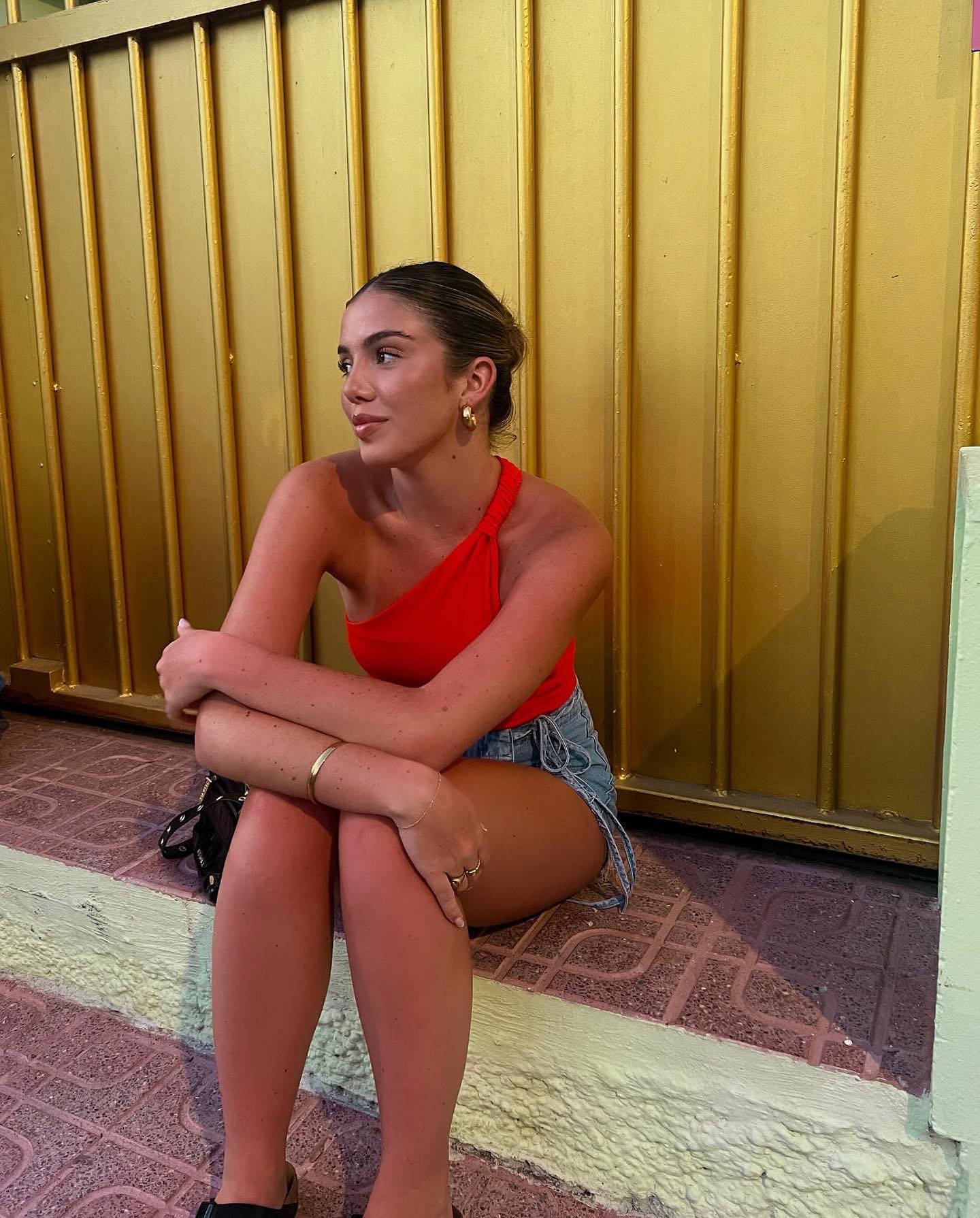 Young adult woman in a red top and jean shorts sits on steps in front of a gold door. She has brown hair in a bun.