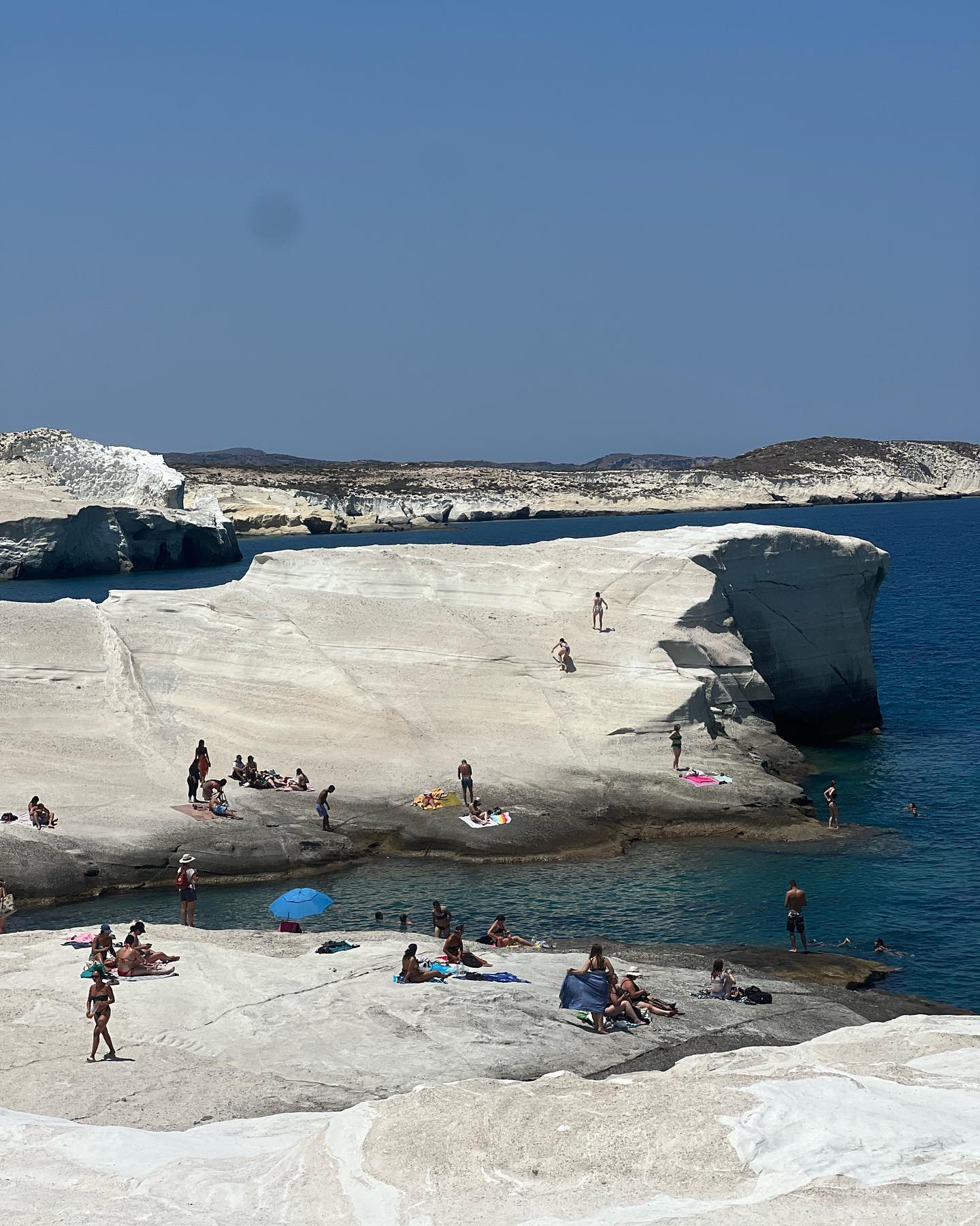 Scenic daytime view of Sarakiniko Beach in Milos, Greece, featuring a white rock landscape, turquoise water, and people relaxing on the rocks.