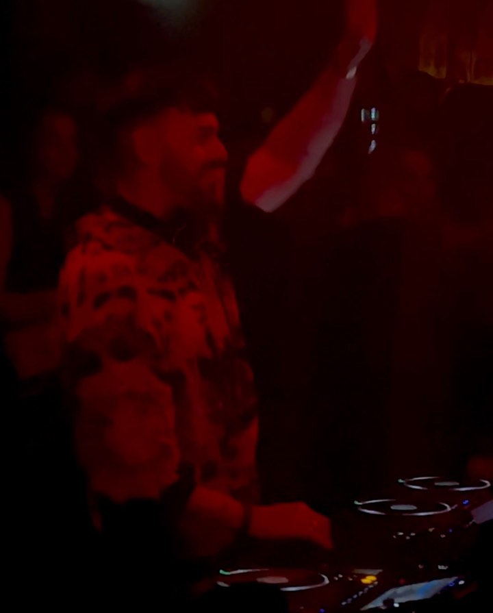 DJ performing in a dimly lit club, wearing a patterned jacket and raising one arm, with a crowd visible in the background.