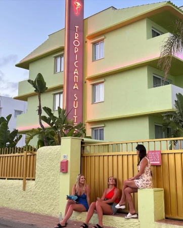 Three women relax on a wall in front of the Tropicana Suites hotel, featuring vibrant colors, palm trees, and a classic Miami vibe.