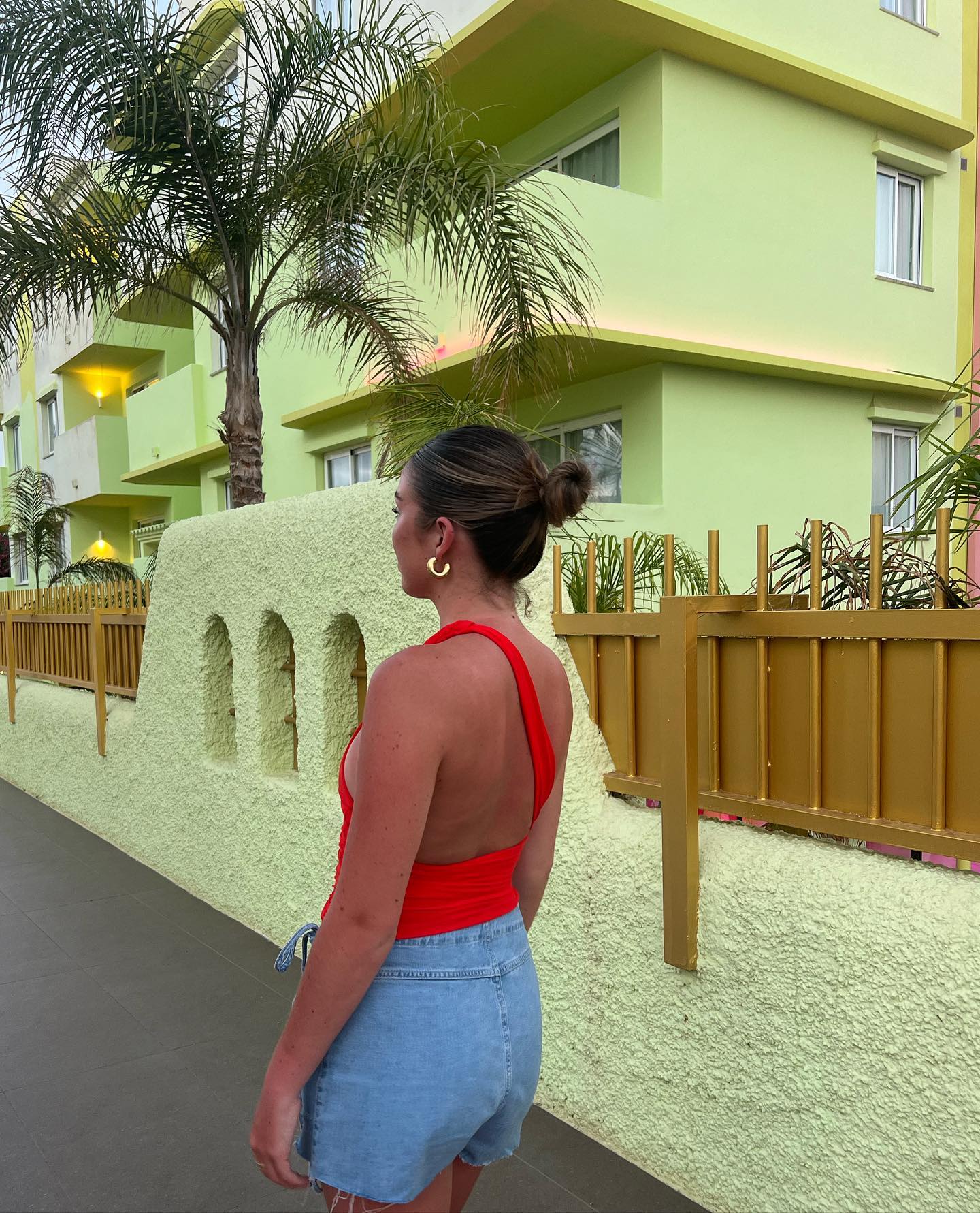 An adult female stands on a sidewalk, wearing a red top and jean shorts, with a light green building and palm tree background.
