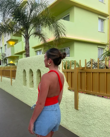 An adult female stands on a sidewalk, wearing a red top and jean shorts, with a light green building and palm tree background.