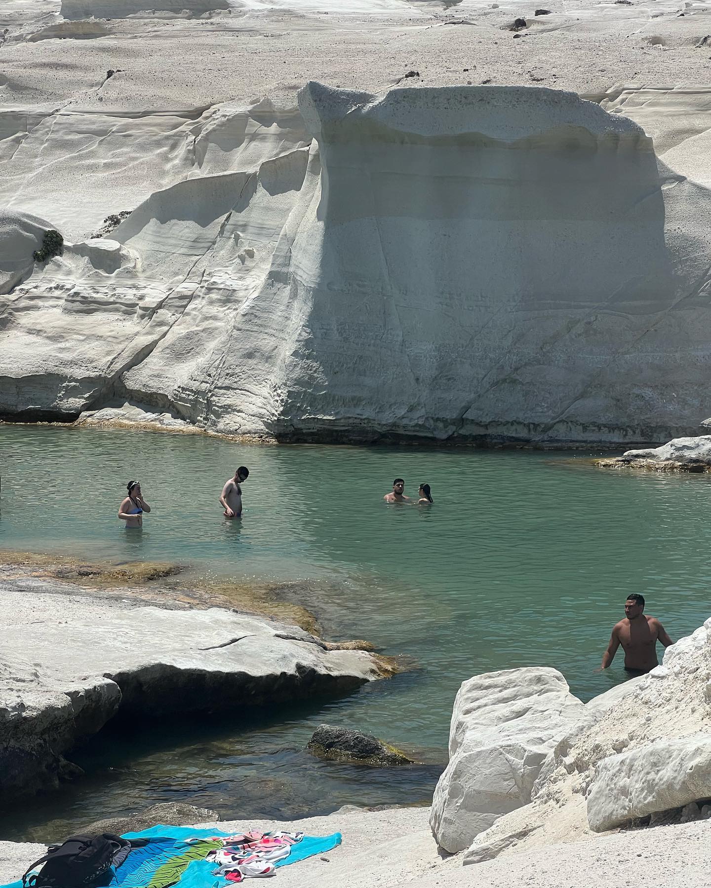 People swim in turquoise water surrounded by white rock formations on a bright day, with beach gear visible in the foreground.