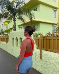 Woman in red top and denim shorts standing outdoors near a pale green building and palm tree