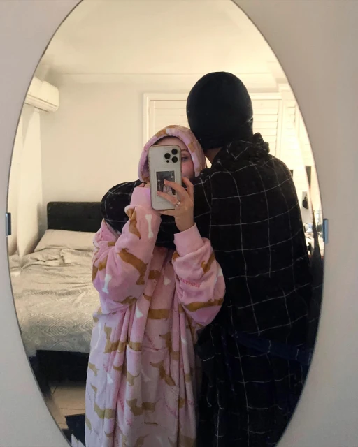 Reflection of a couple in a mirror, one person wearing a pink hooded robe with a dog pattern, the other in a black robe and beanie.