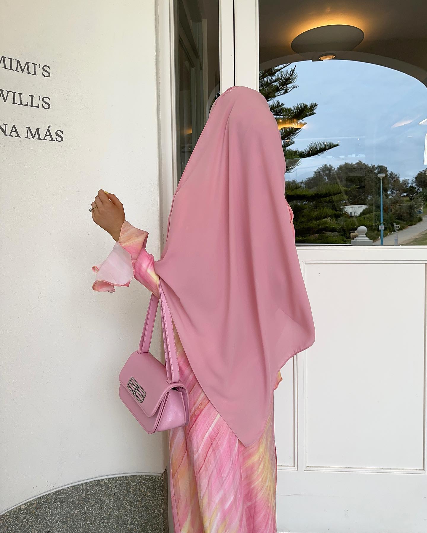 Woman in pink hijab and matching dress poses with a pink handbag in front of a white wall, creating a fashionable, monochromatic look.