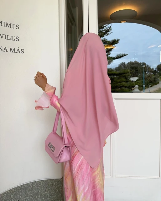 Woman in pink hijab and matching dress poses with a pink handbag in front of a white wall, creating a fashionable, monochromatic look.