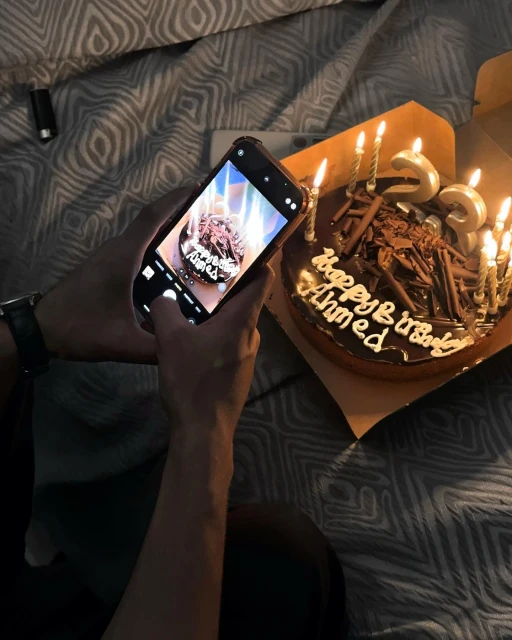 Person takes a picture with their cellphone of a chocolate birthday cake with lit candles on a bed with patterned gray bedding.