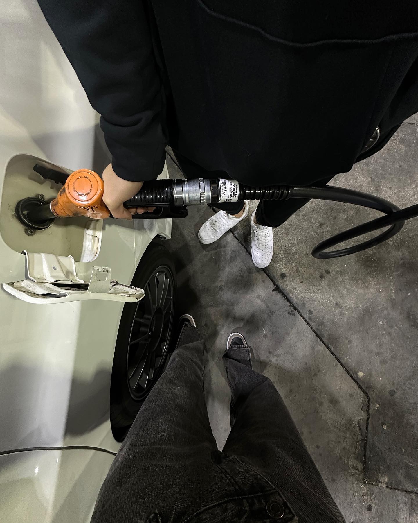 Two people at a gas station are filling up a white car with fuel using an orange nozzle and a black hose on a concrete surface.