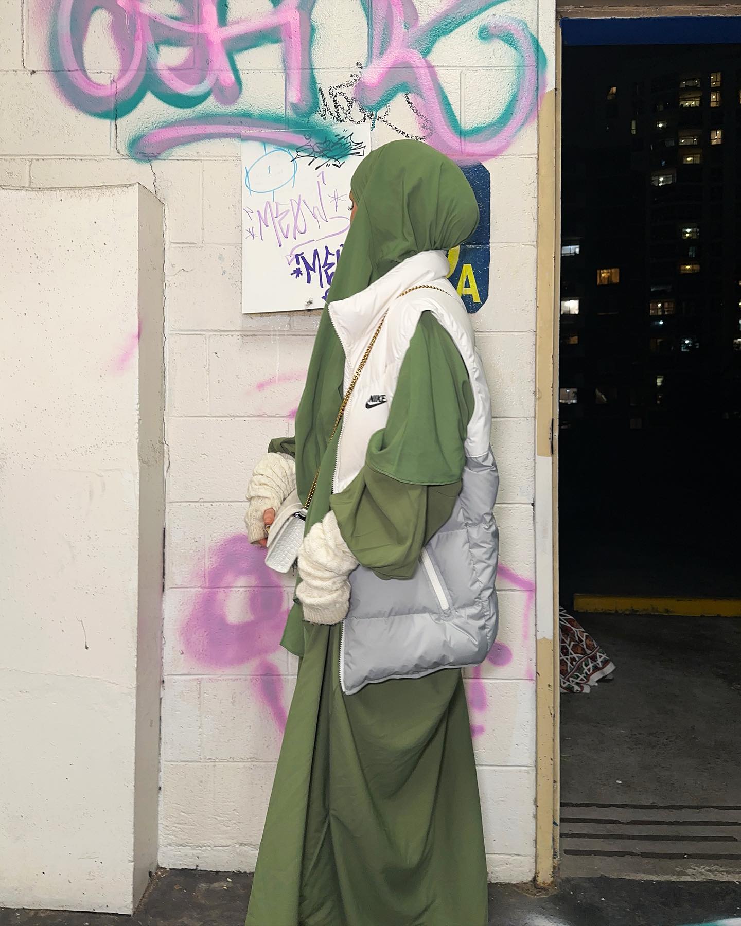 Woman wearing a green hijab, puffer vest, and long dress poses outdoors near a graffiti-covered wall and a dark parking garage entrance.