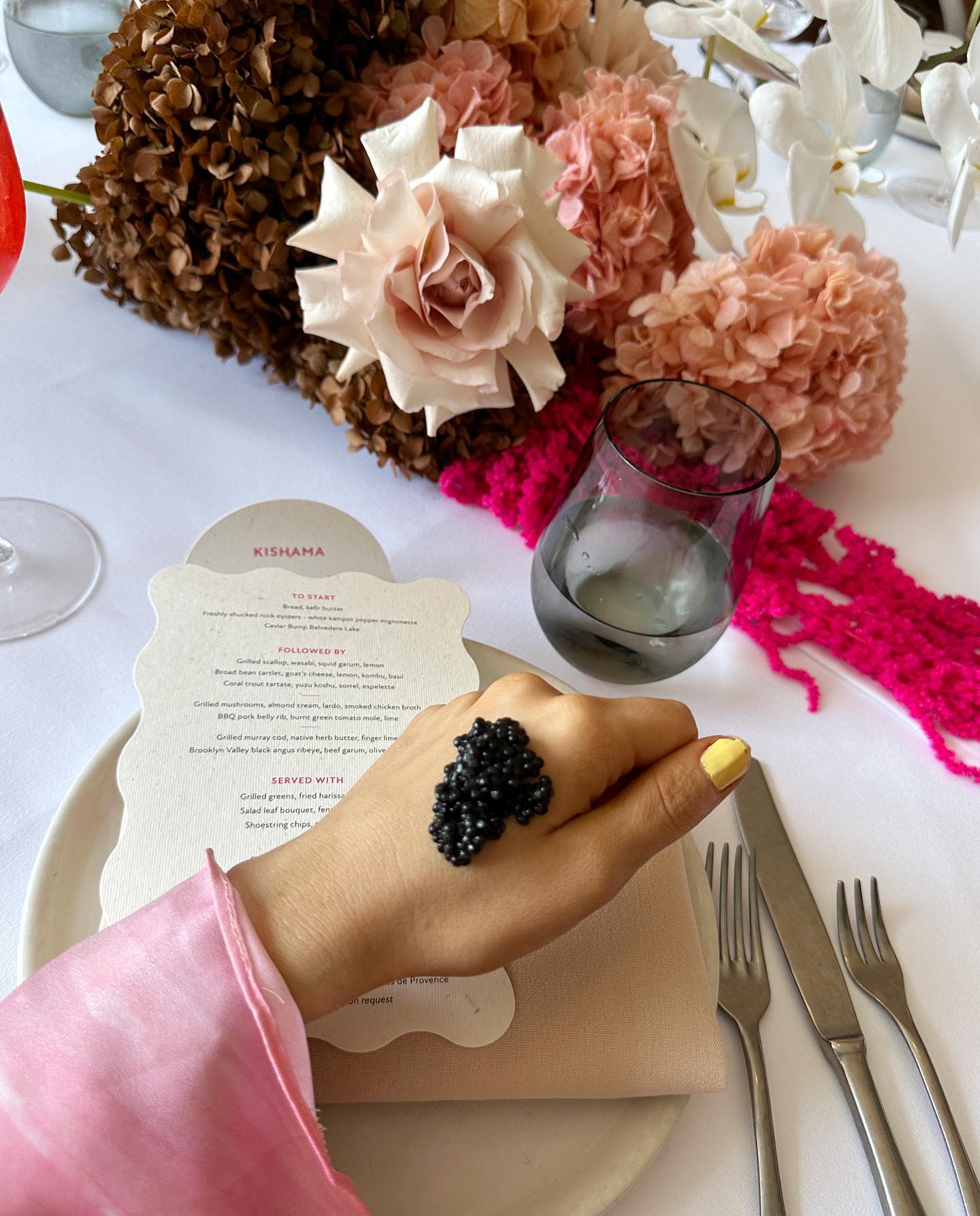 Caviar on a woman's hand at a dining table with a menu, glass of water, silverware, and floral arrangement.