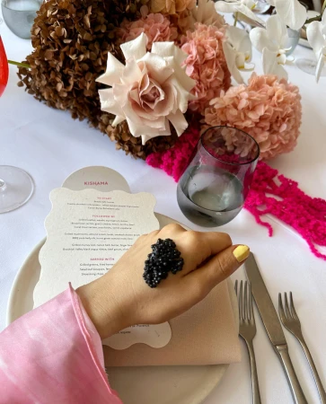 Caviar on a woman's hand at a dining table with a menu, glass of water, silverware, and floral arrangement.