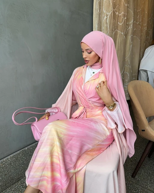 A woman in a pink hijab and tie-dye dress sits indoors, holding a pink purse, wearing a watch and rings, with a gray wall behind her.