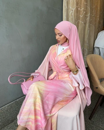 A woman in a pink hijab and tie-dye dress sits indoors, holding a pink purse, wearing a watch and rings, with a gray wall behind her.