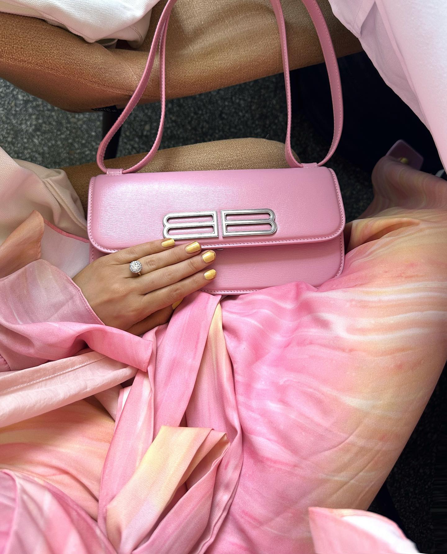 A pink Balenciaga purse sits on a person's lap, with yellow nails and a diamond ring visible. The dress is pink and peach.