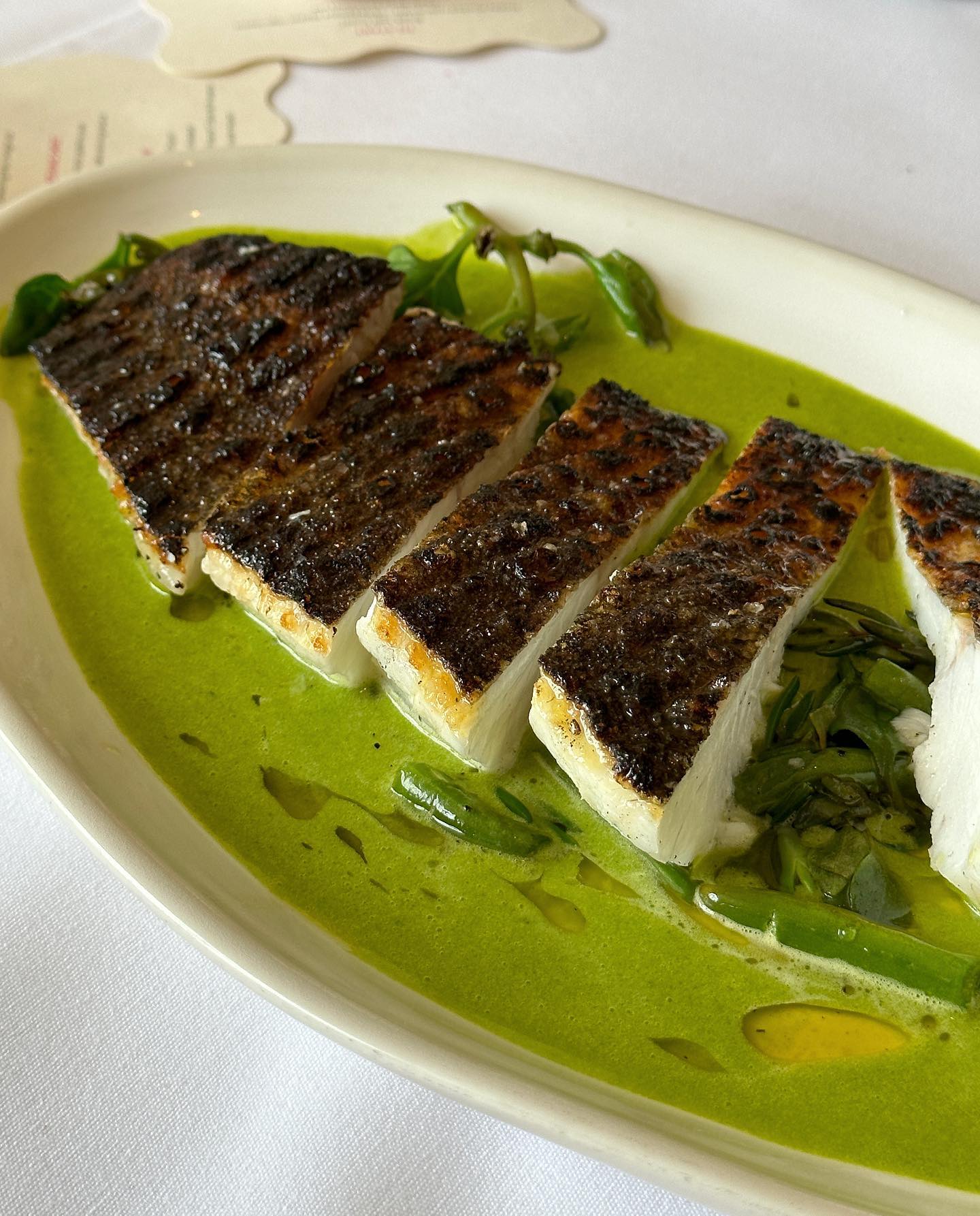 Close-up of sliced, grilled fish served on a white plate with green sauce and herbs, showcasing culinary presentation and food photography.