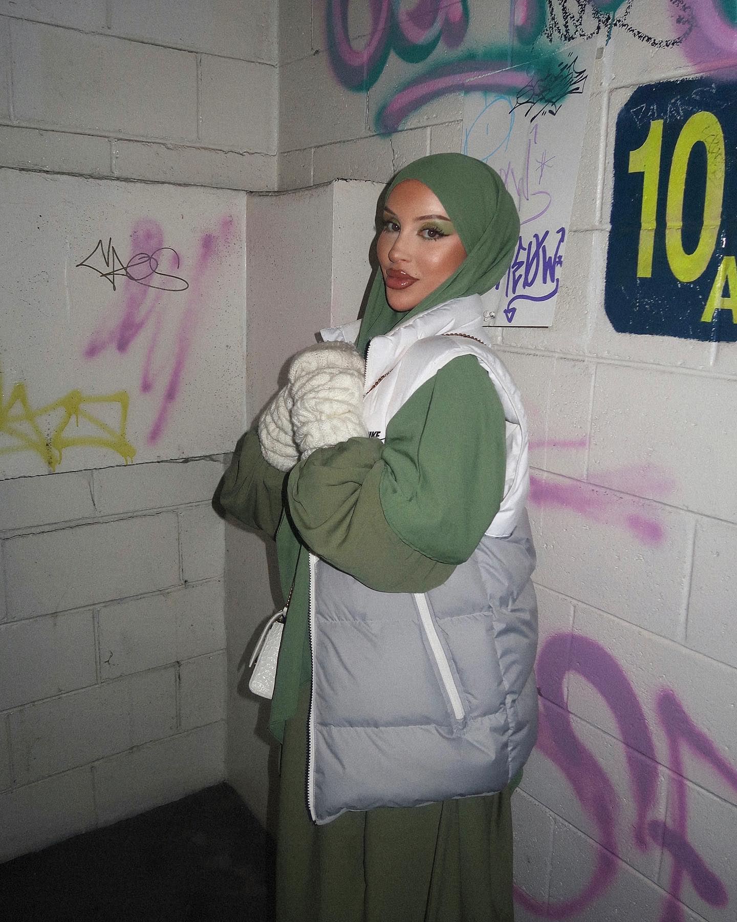 Woman in headscarf and vest stands against graffiti wall in urban setting. The photo features olive green and white colours.