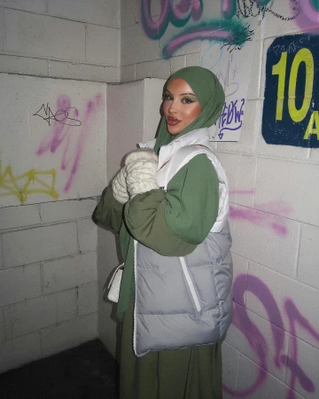 Woman in headscarf and vest stands against graffiti wall in urban setting. The photo features olive green and white colours.