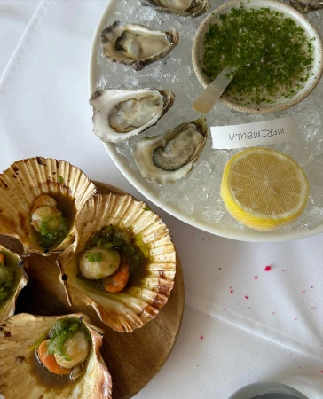 Overhead shot of scallops in shells on a wood board and oysters on crushed ice with lemon and green sauce on a white tablecloth.