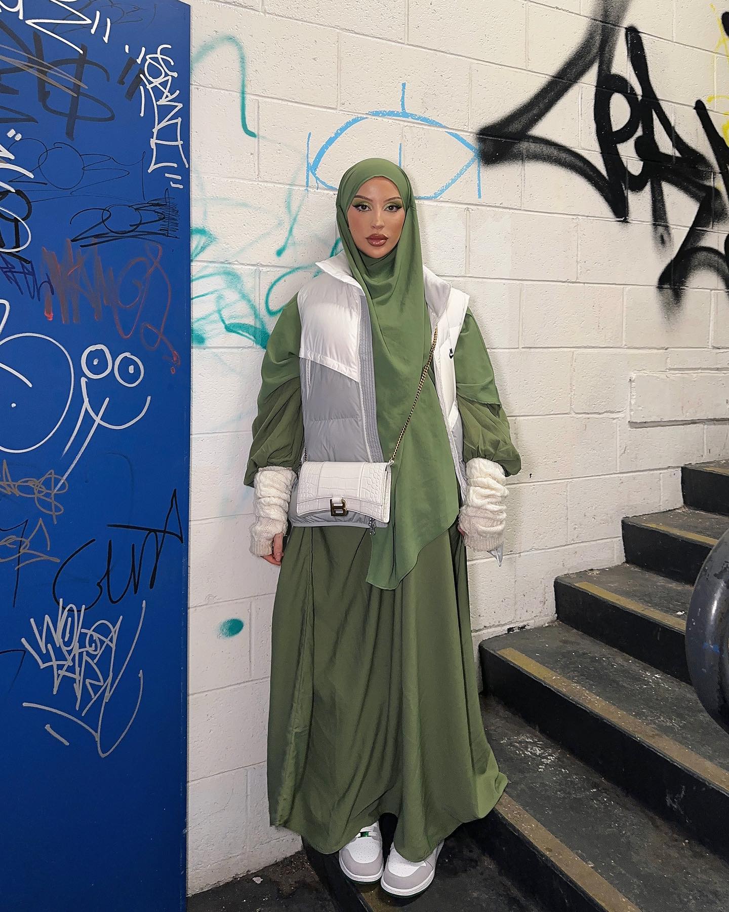 Woman in hijab, long green dress, and white puffer vest poses against graffiti wall with a small purse and grey and white sneakers.