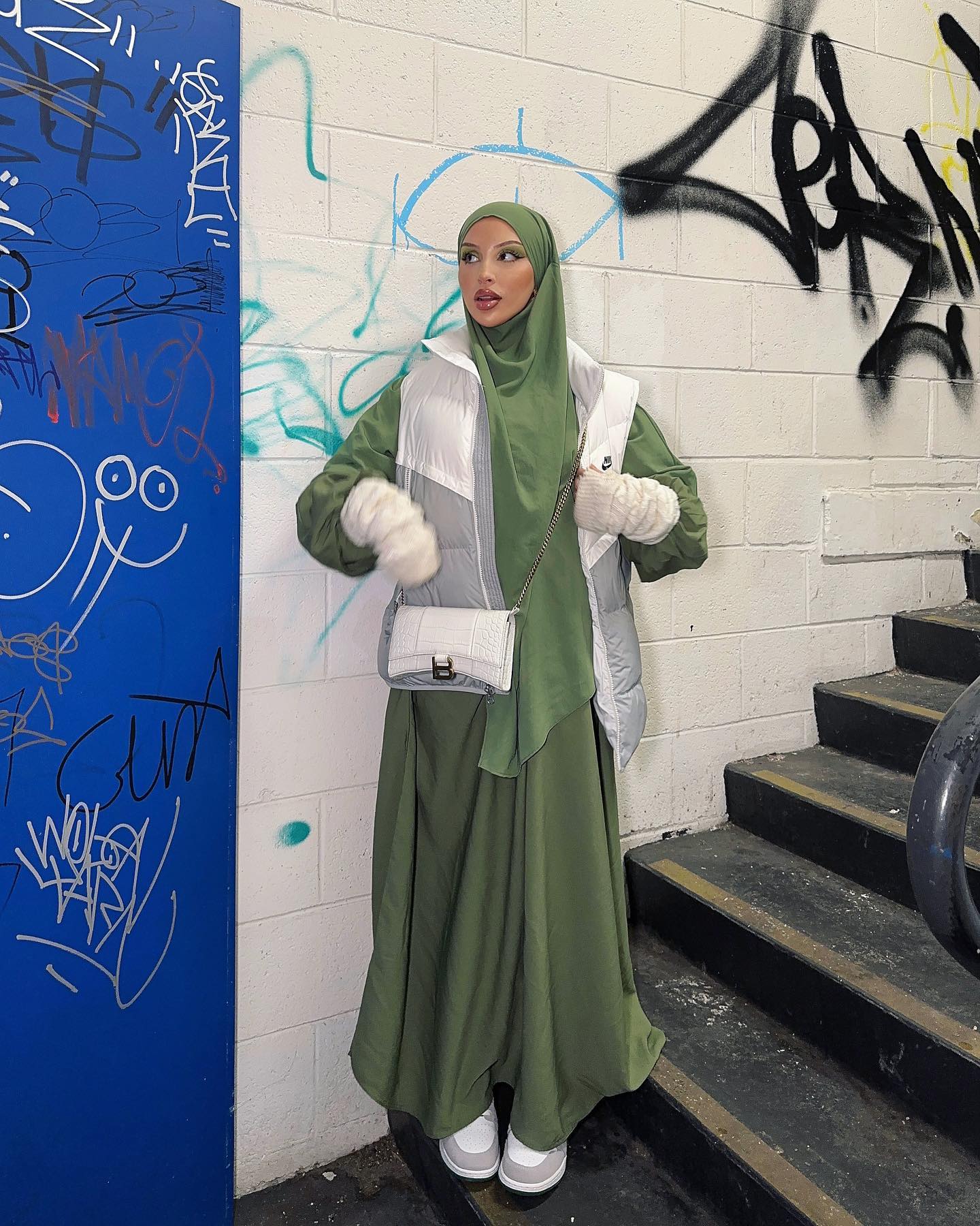 A stylish adult female in an olive-green dress and hijab, standing against a graffiti wall, accessorized with a white vest, bag, and gloves.