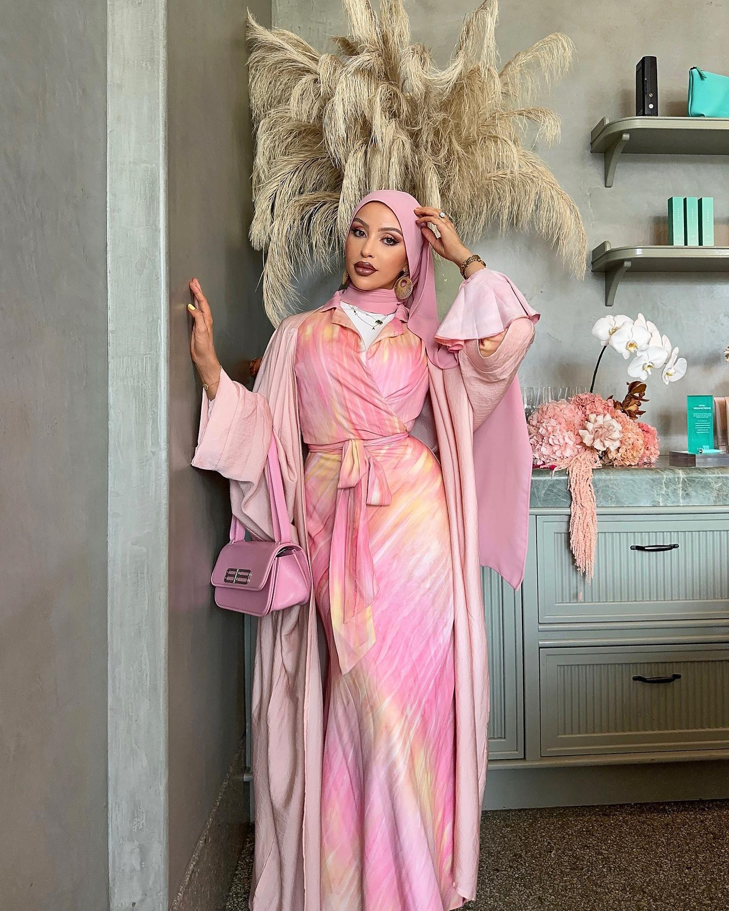 Woman wearing pink hijab, tie-dye wrap dress, and carrying a pink purse posing in an indoor environment with neutral wall and decor.