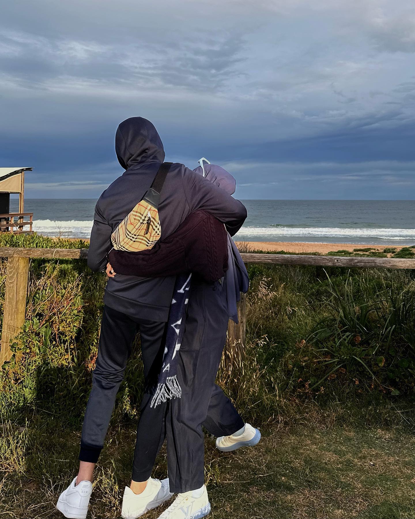 Two figures embracing on a dune overlook, with a hooded person wearing a cross-body bag and another in a scarf, set against a beach and cloudy sky.