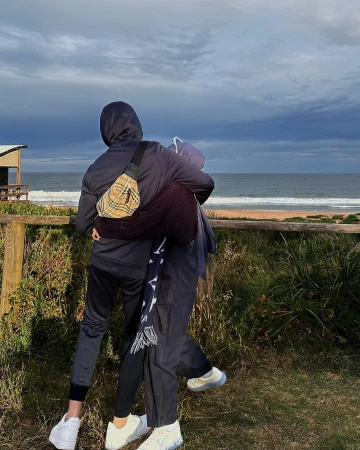 Two figures embracing on a dune overlook, with a hooded person wearing a cross-body bag and another in a scarf, set against a beach and cloudy sky.
