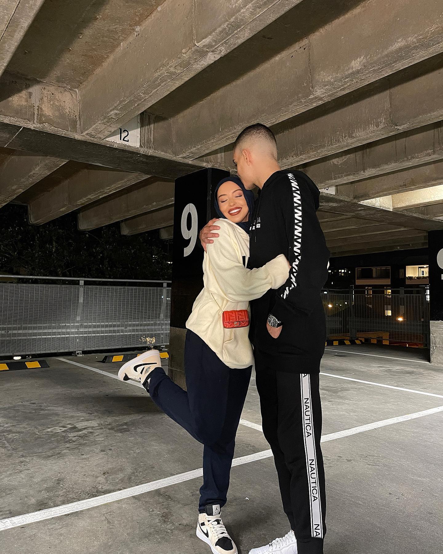 A happy couple embraces in a parking garage: A woman in a hijab and cream-colored sweatshirt hugs a man in a black hoodie and sweatpants.
