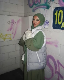 Content creator Kishama Meridian wearing a green hijab and white puffer vest against a graffiti-tagged brick wall in Sydney.