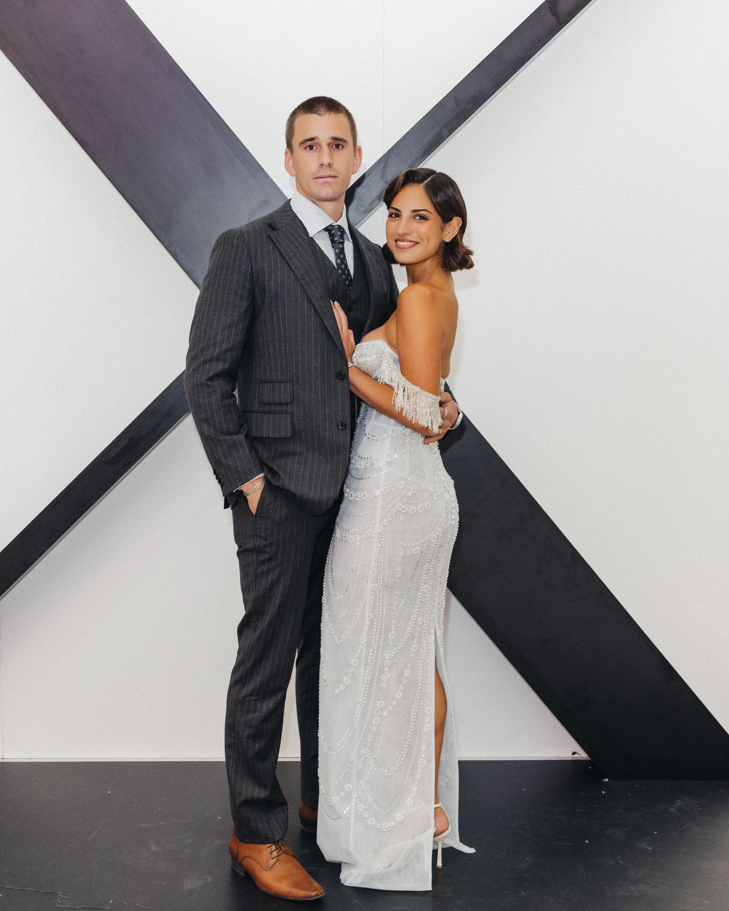 Elegant couple poses in front of a monochrome geometric backdrop. The man wears a pinstripe suit, while the woman stuns in a beaded gown.