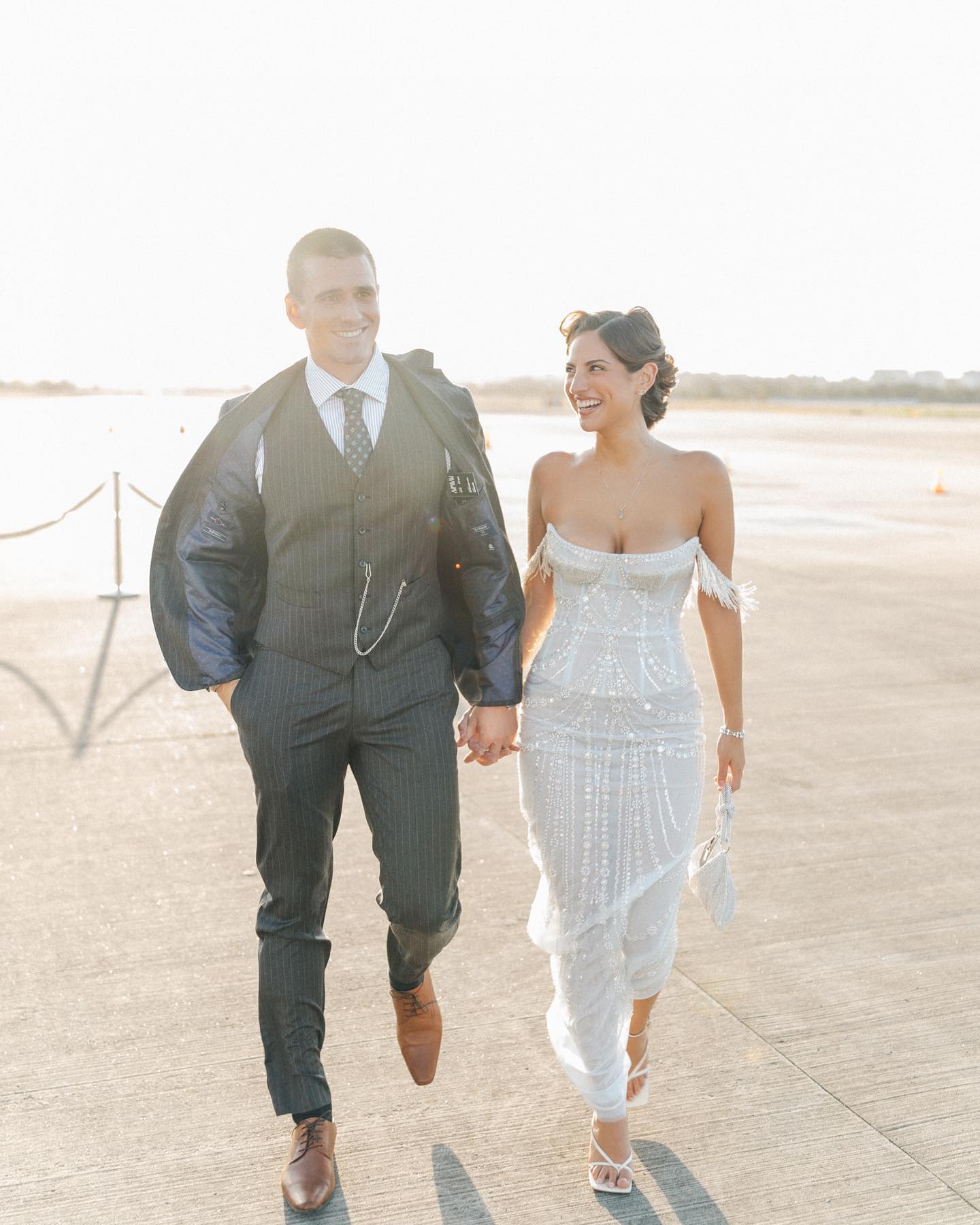 Smiling couple in wedding attire holding hands and walking on an open field during daytime, with the man carrying his suit jacket.