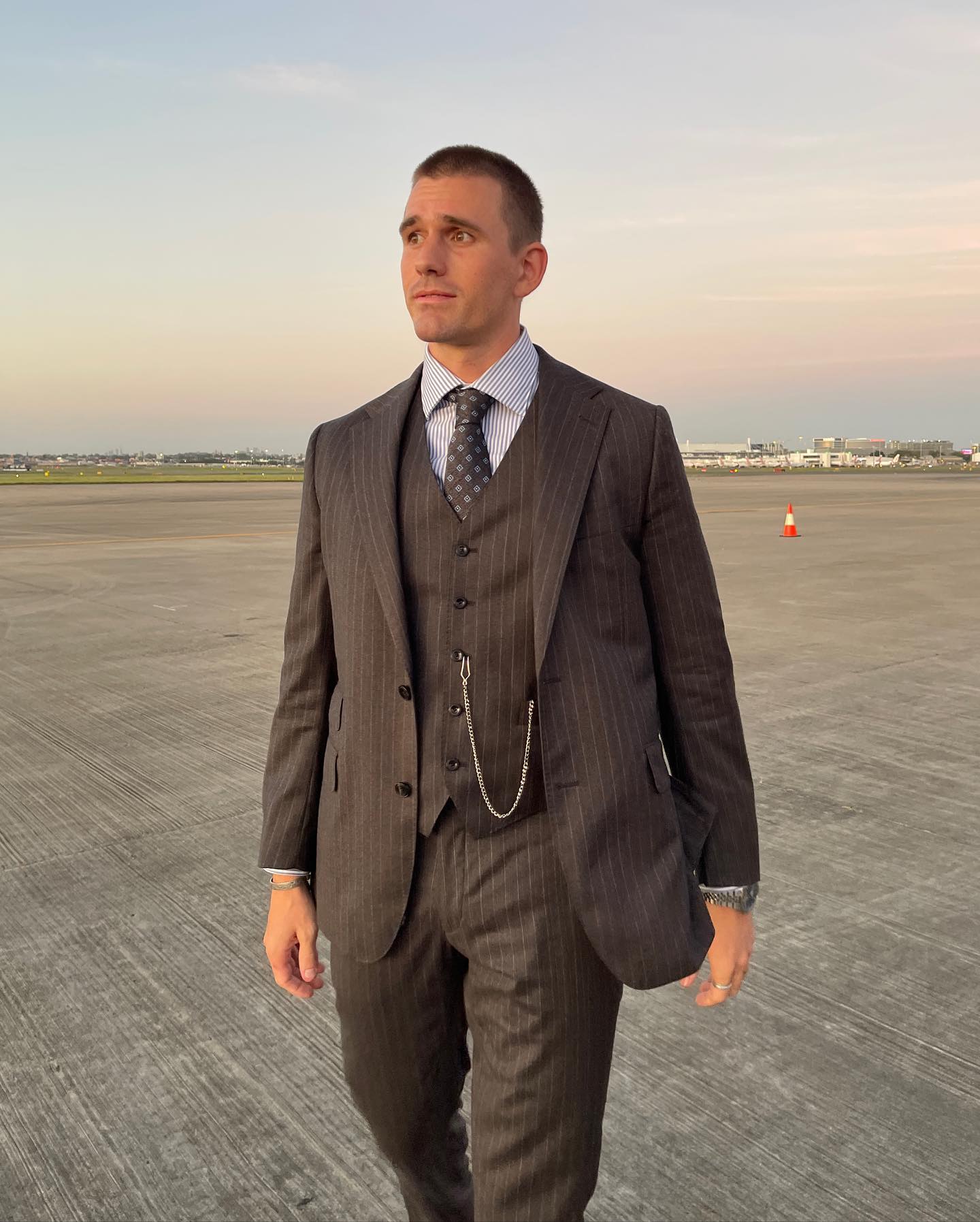 Man in suit with vest and tie walks on an airport runway under an open sky at dusk wearing a suit and dress shoes.