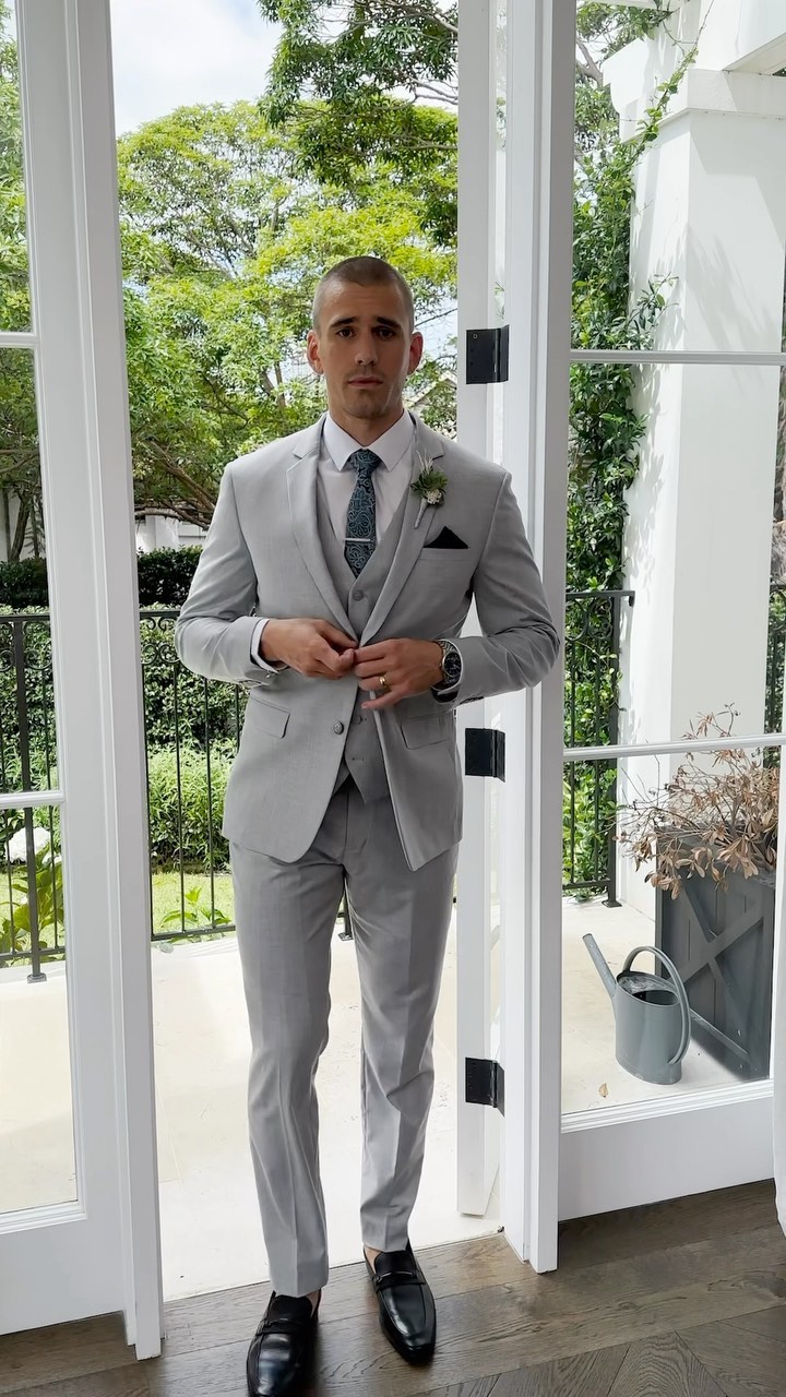 Man in a grey suit poses confidently by a white-framed doorway with outdoor foliage backdrop; elegant formal wear fashion.