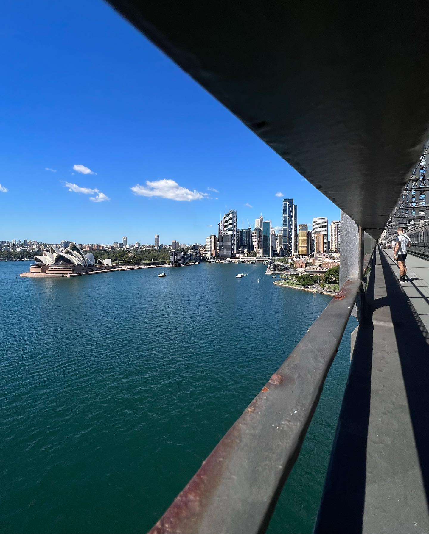 Sydney Harbour with the Opera House and city skyline in the background, seen from the Sydney Harbour Bridge, with a person walking.
