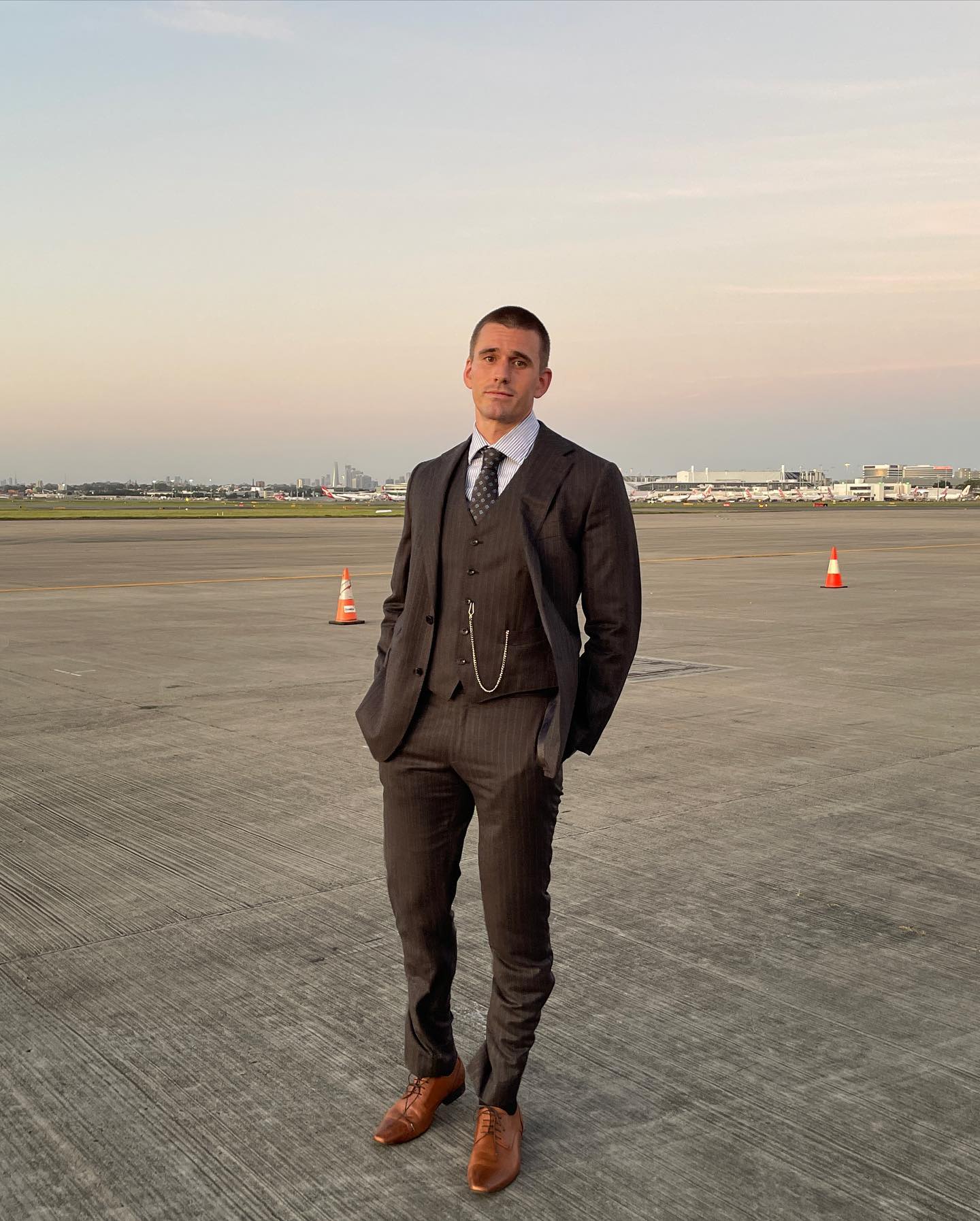 Full-length portrait of a man in a three-piece suit outdoors on an airport tarmac under a blue sky with wispy clouds.