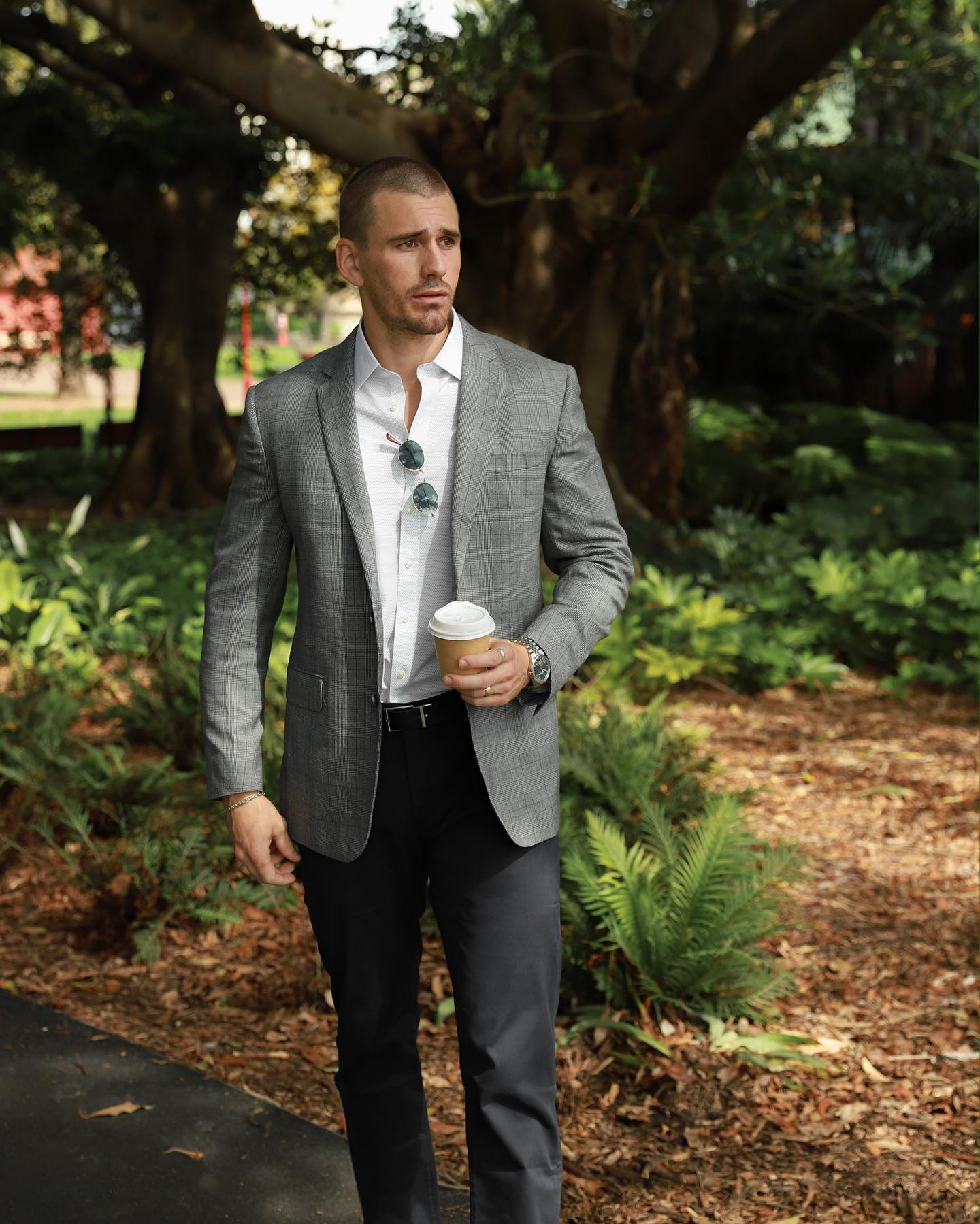 Man in grey blazer and dark pants holding a coffee cup in a park setting with leafy trees and ferns, captured in medium shot.