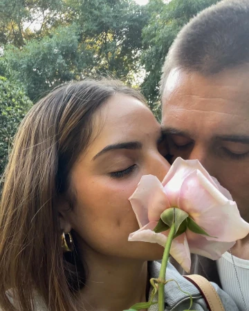 Close-up portrait of a woman and man with eyes closed, gently sniffing a large pale pink rose held between them.