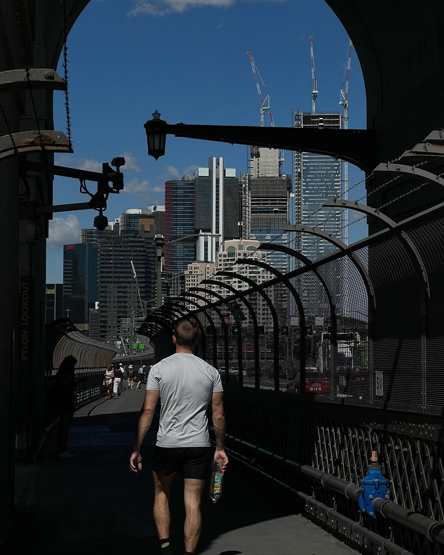 Man walks on bridge with modern city skyline in background, Sydney Harbour Bridge visible, bright daylight, blue sky, urban scene.