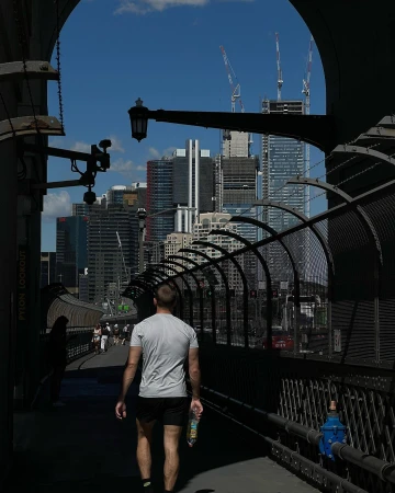 Man walks on bridge with modern city skyline in background, Sydney Harbour Bridge visible, bright daylight, blue sky, urban scene.